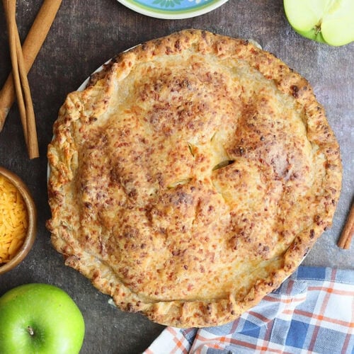 Baked Granny Smith apple pie with cheddar cheese crust with a plate, fork, cinnamon sticks, shredded cheese, and green apples off to the side.