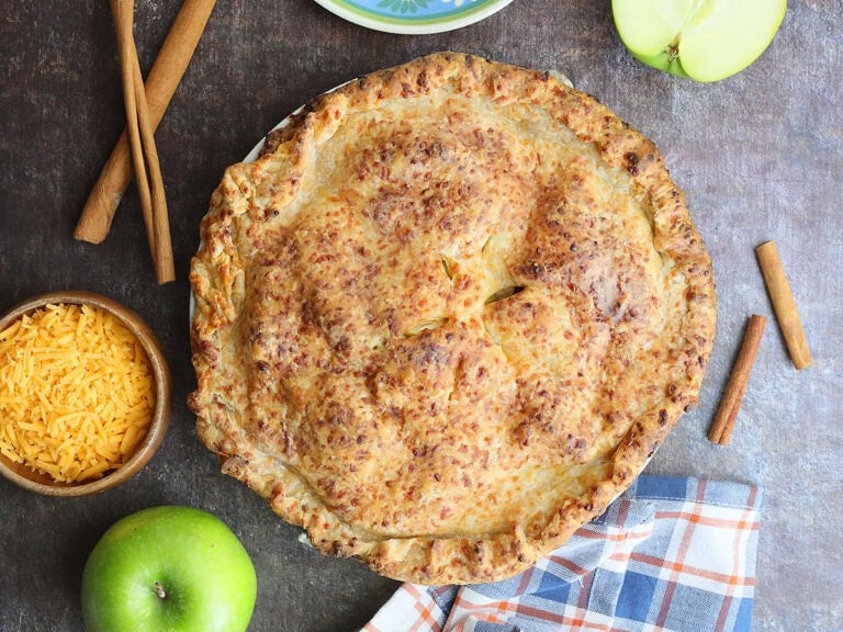 Baked Granny Smith apple pie with cheddar cheese crust with a plate, fork, cinnamon sticks, shredded cheese, and green apples off to the side.