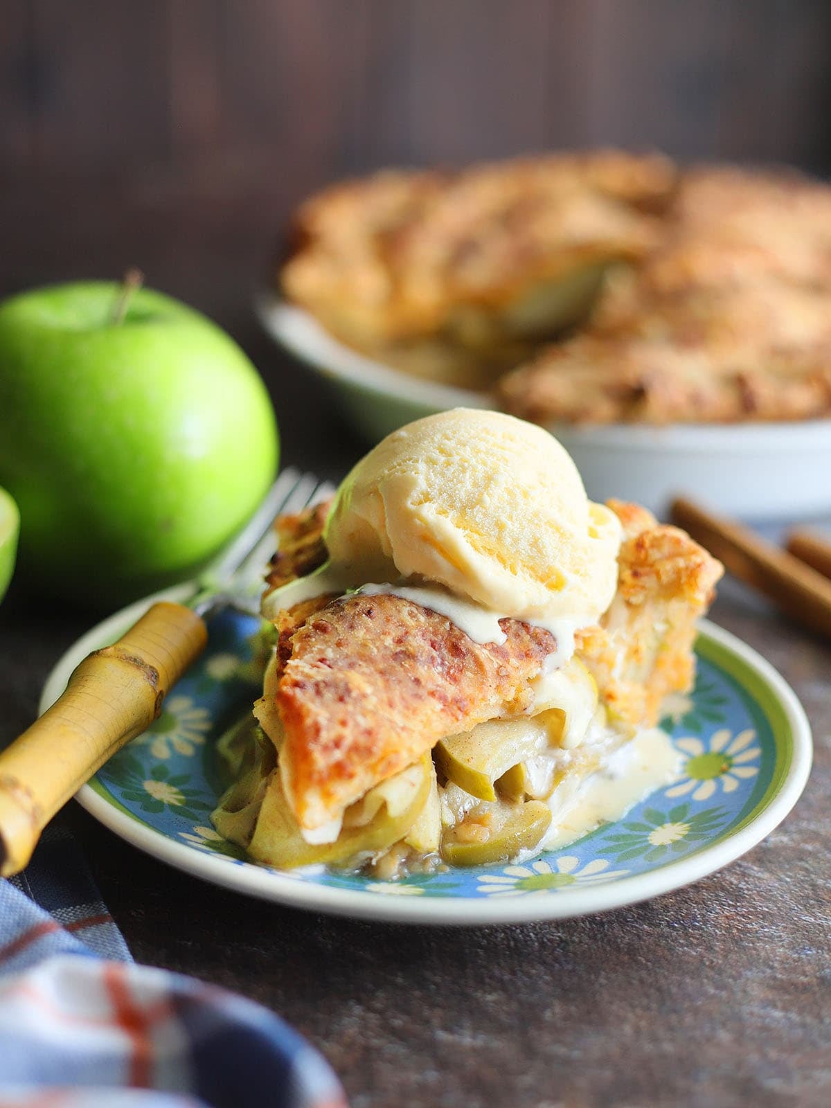 A slice of Granny Smith apple pie with cheddar cheese crust on a plate with a scoop of vanilla ice cream.