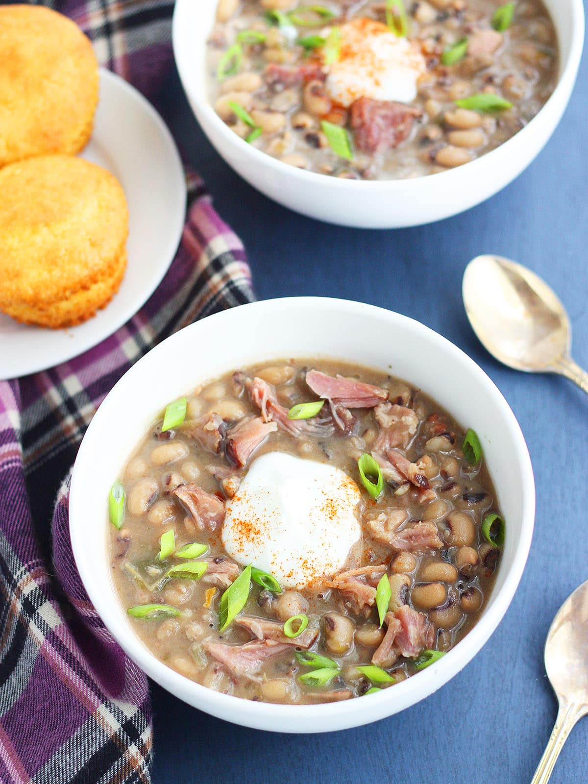 Two bowls of of black eyed pea soup garnished with a dollop of sour cream and cayenne pepper with a plate of corn muffins on the side.