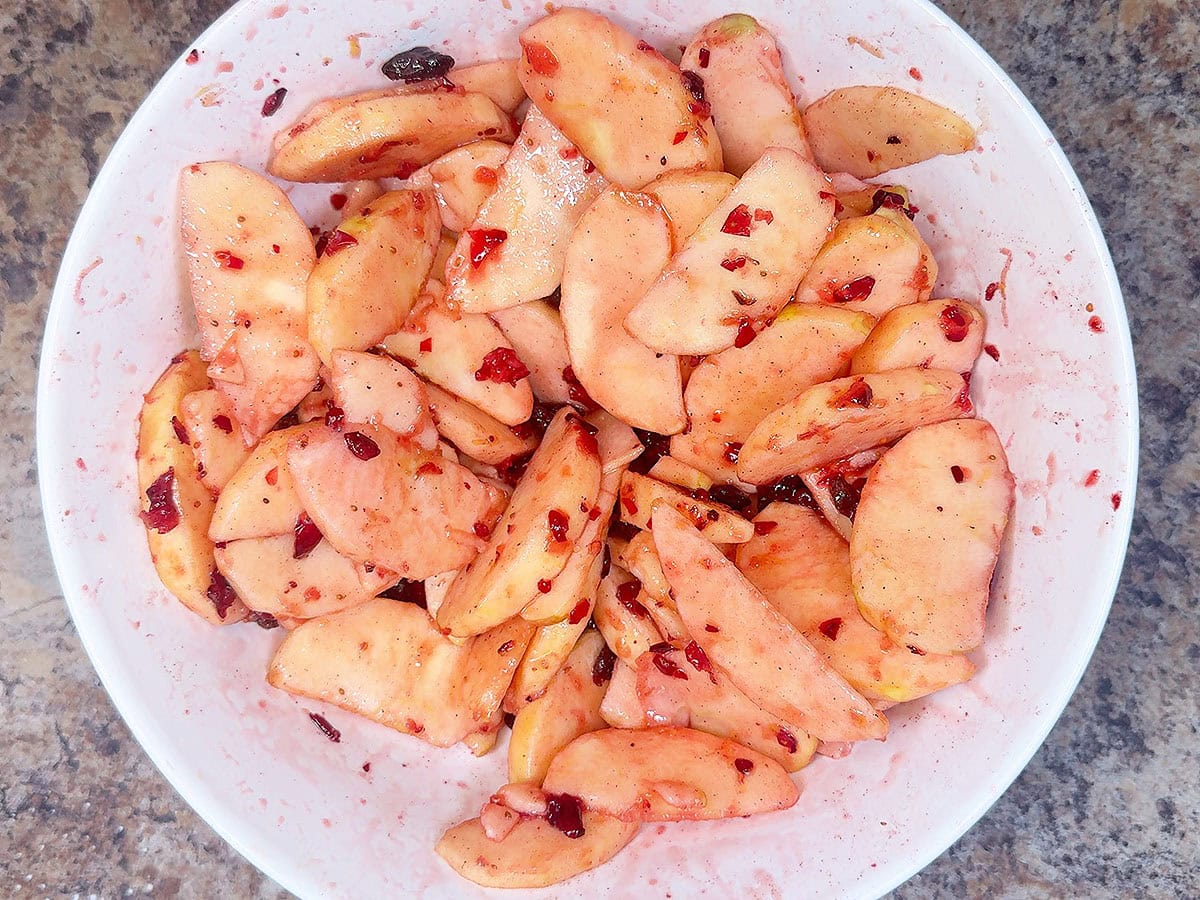 Sliced Fuji apples mixed with cinnamon, sugar, and chopped cranberries in a mixing bowl.