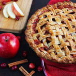 Cranberry apple pie with a golden lattice crust. Whole and sliced apples, fresh cranberries, and cinnamon sticks are arranged around the pie.