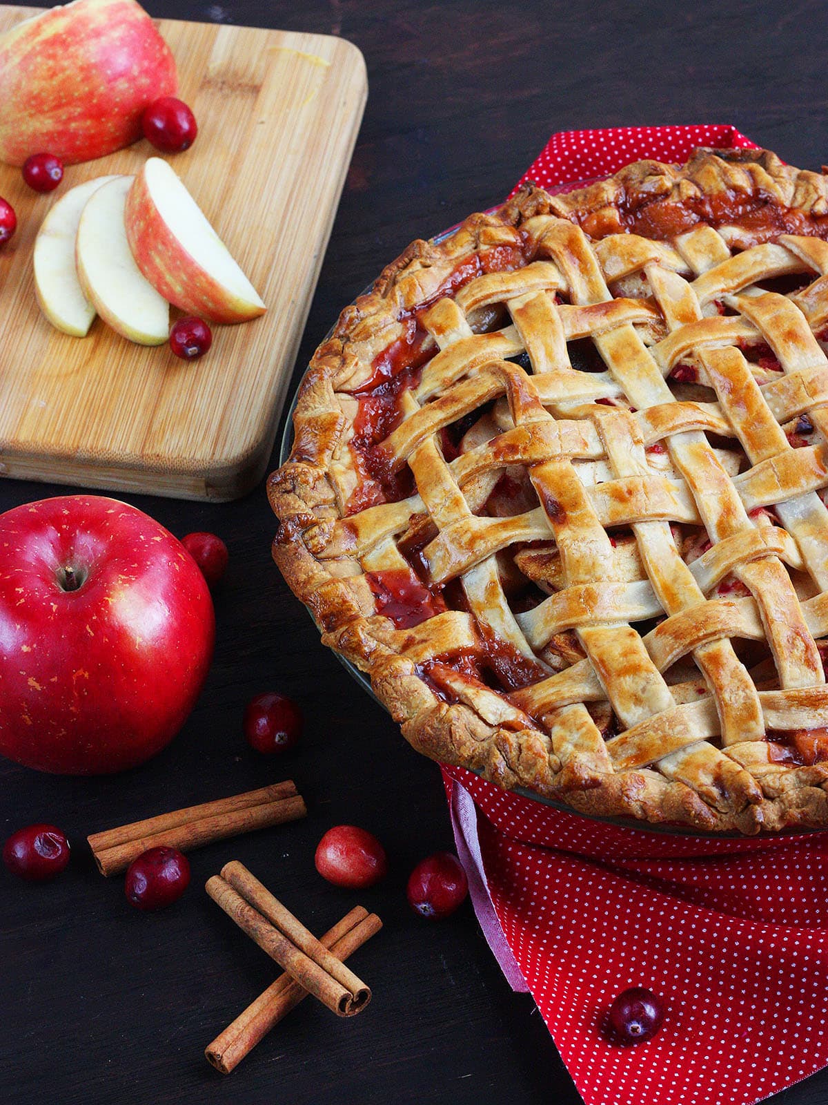 Cranberry apple pie with a golden lattice crust. Whole and sliced apples, fresh cranberries, and cinnamon sticks are arranged around the pie.