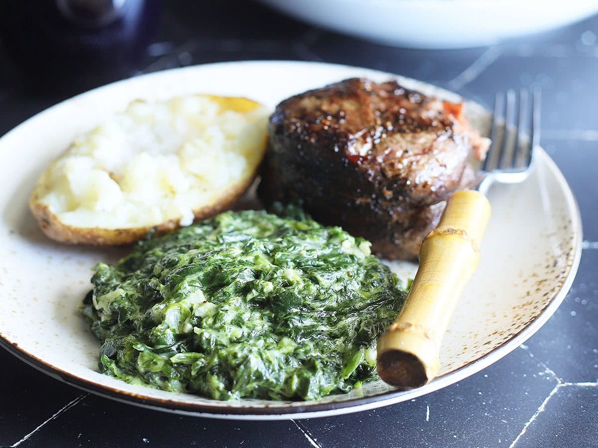 Serving of creamed spinach on a plate with a grilled steak and baked potato.