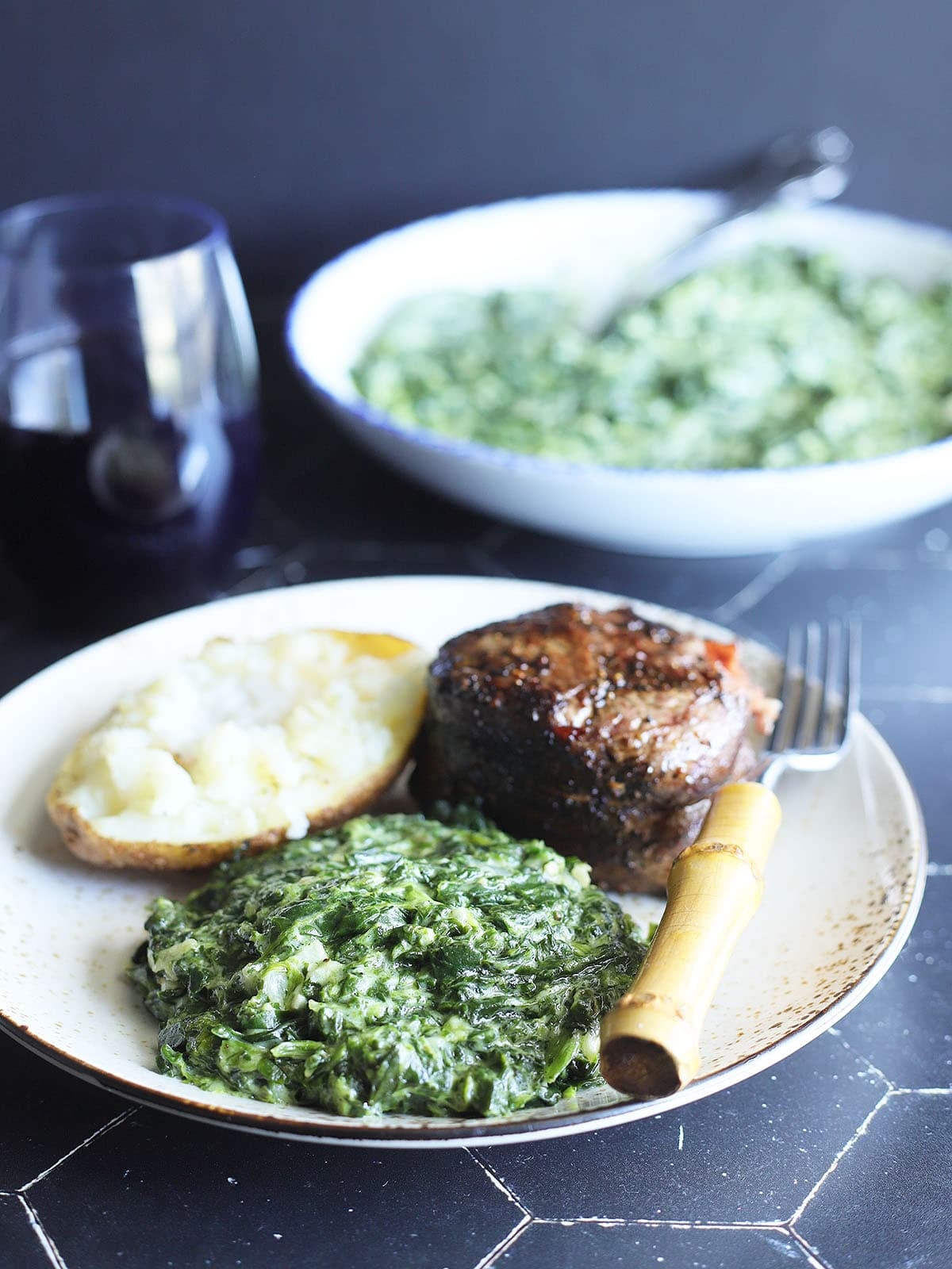 Serving of creamed spinach on a plate with a grilled steak and baked potato.